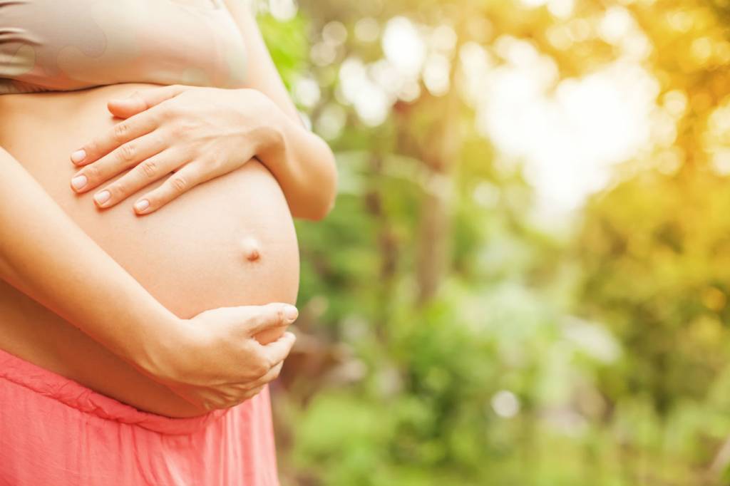 Barriga de mulher grávida de pele branca. Ela está com os dois braços sobre a barriga, que está desnuda.