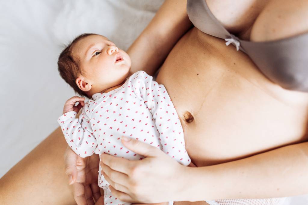 bebê recém-nascido de macacão no colo da mãe, que está usando lengerie. Ambos têm pele clara. O bebê está olhando para a mãe.