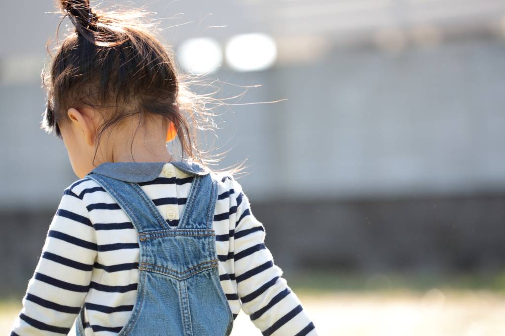 criança pequena de costas, com o cabelo castanho prero, usando camisa listrada e jardineira jeans