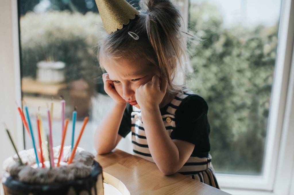 Menina triste diante do bolo de aniversário. A garota tem o cabelo preso, usa um chapéu de papelão na cabela e veste uma camiseta preta de mangas curtas com um vestido listrado preto e branco. Há velas coloridas sobre o bolo.