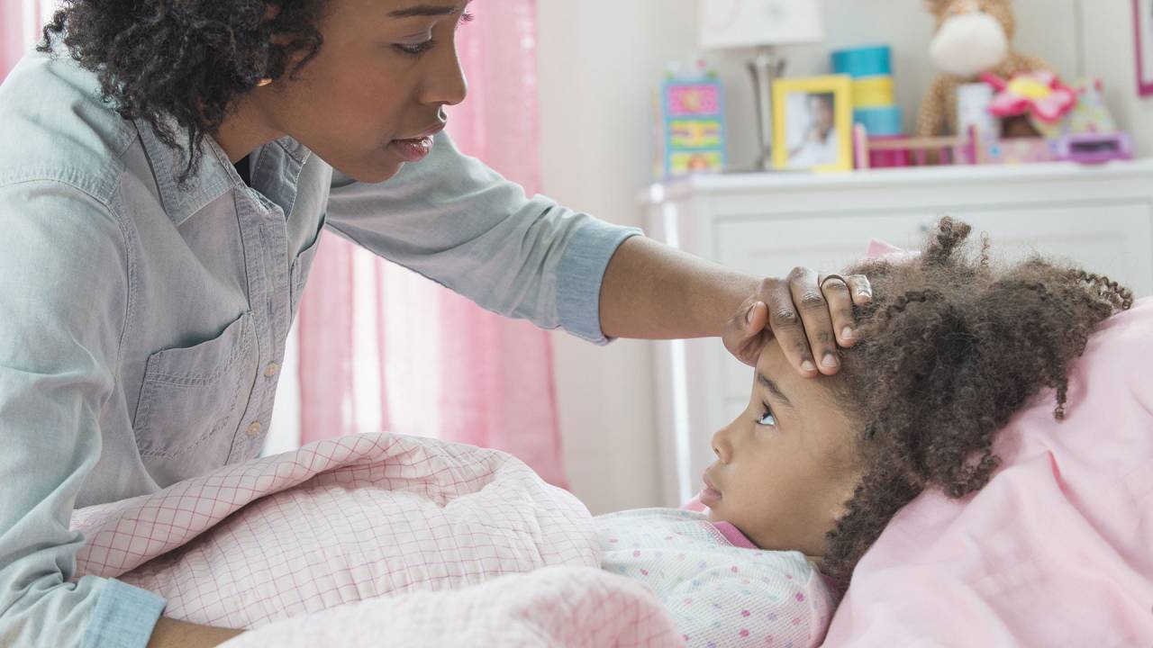 mãe com a mão na testa da filha criança, que está deitada em uma cama
