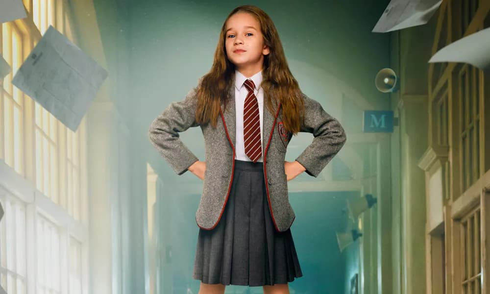 Cena de Matilda o musical. Uma menina vestida com uniforme tradicional americano, de saia, gravata, camisa e terninho, está com as mãos na cintura.