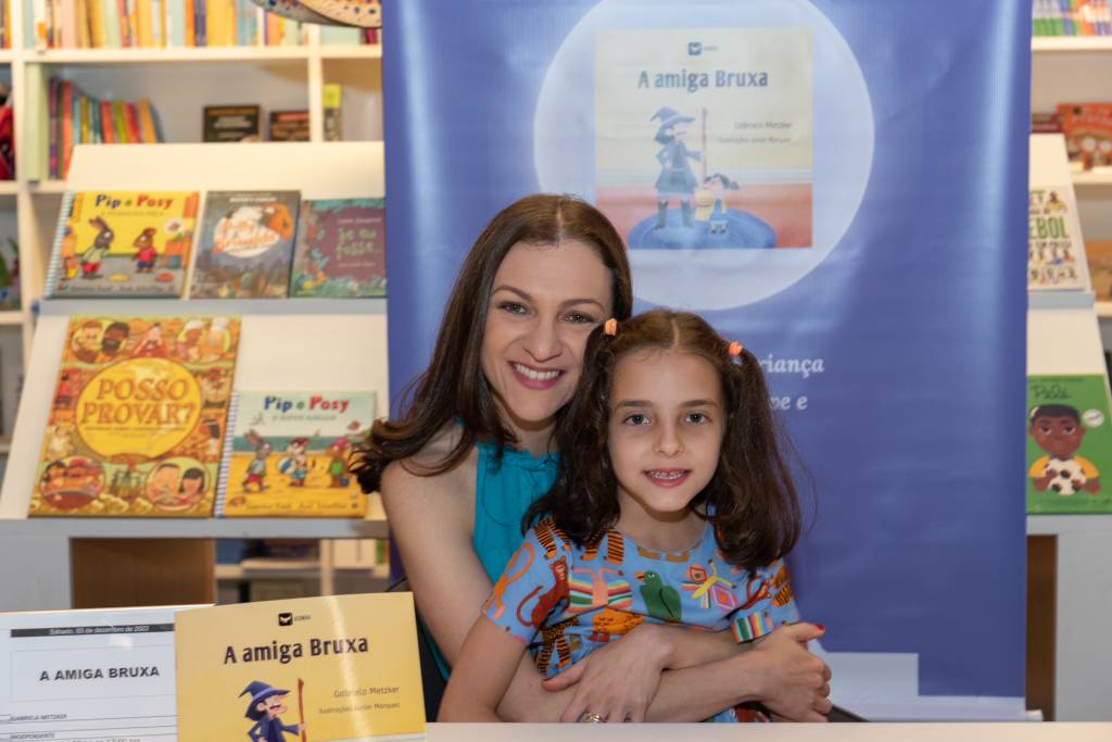 gabriela metzker autora Gabriela Metzker, autora do livro, abraçando a filha no lançamento do livro. As duas estão sorrindo. Sobre a mesa, está o livro A Amiga Bruxa.
