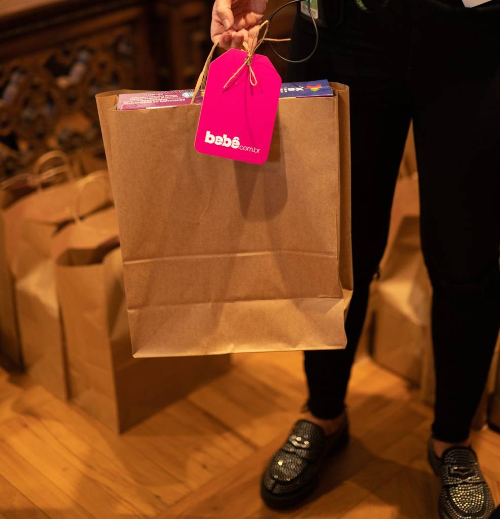 Presentes especiais casa clã Mulher vestida de preto segurando sacola de papel com tag pink escrito Bebê.com.br