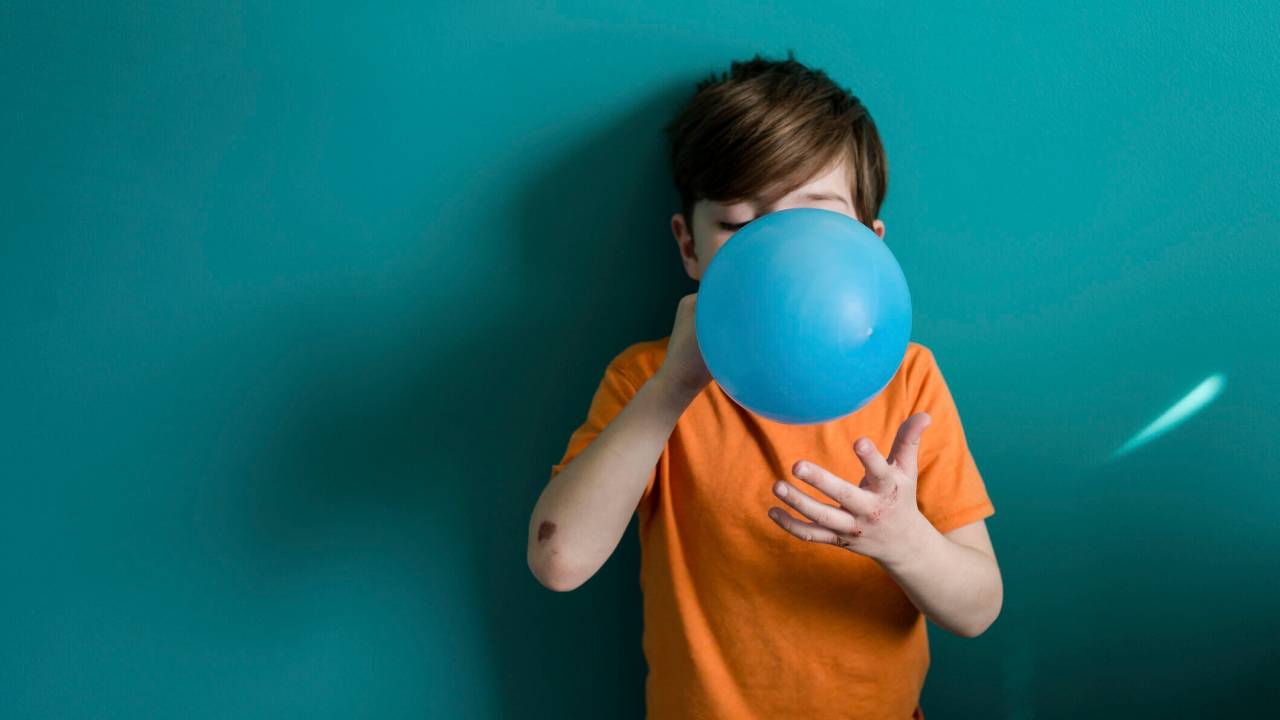 Menino, por volta de 5 anos, branco, usando camiseta laranja e enchendo uma bexiga azul. Tem cabelo castanho escuro.