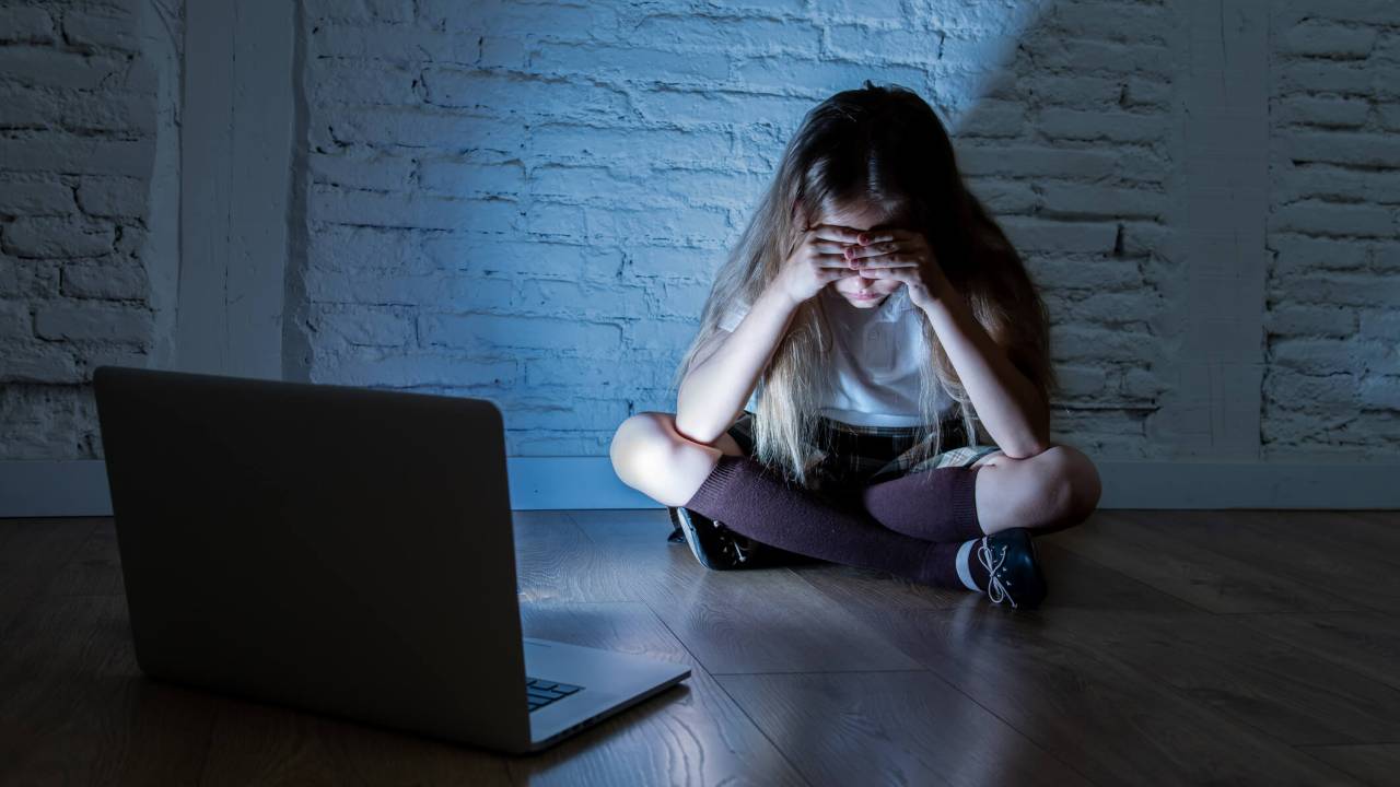 Criança sentada no chão, em frente a um notebook, no escuro. Ela está com a cabeça baixa e o rosto coberto pelas mãos.