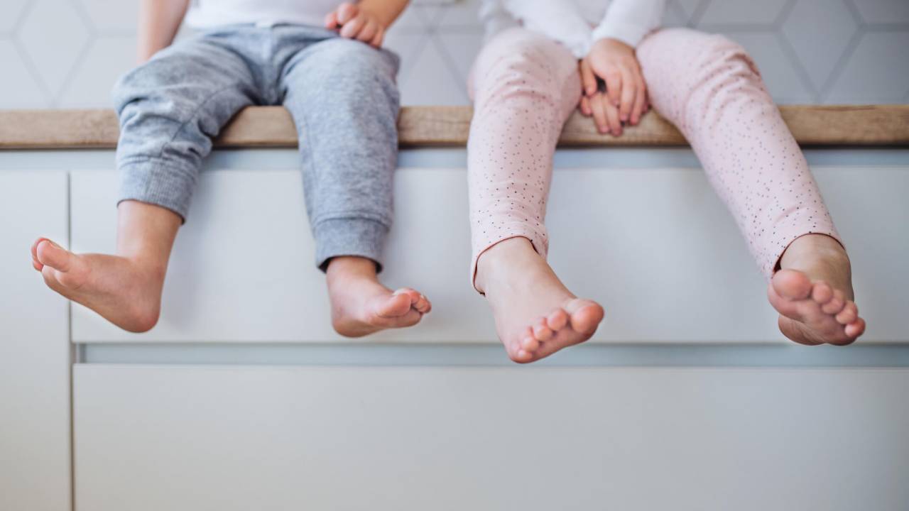 Duas crianças descalças, sentadas sobre uma cômoda. Elas estão de camiseta branca e calça comprida - uma cinza e outra rosa. Não é possível ver os rostos.