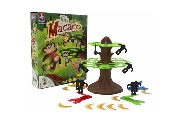 de um lado, a caixa de papelão que é a embalagem do jogo Pula Macaco. Ao lado, o jogo aberto, com uma árvore de plástico, macacos também de plásticos pendurados nela e bananas de papel no chão