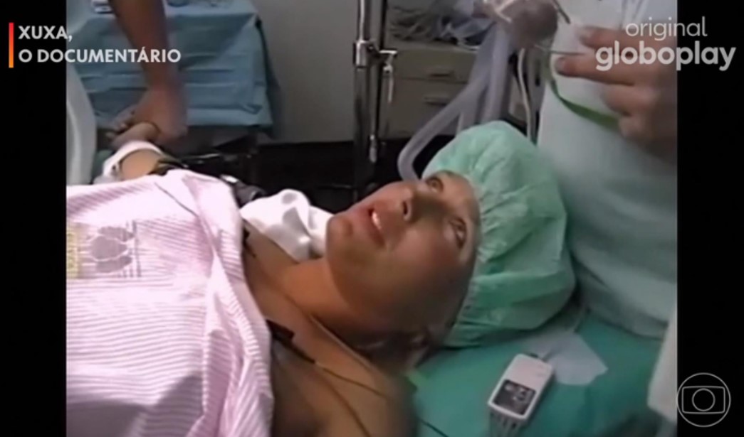 Imagem de Xuxa em parto da Sasha. Ela está deitada na mesa de cirurgia, com touca verde e coberta por lençol branco, olhando para cima.