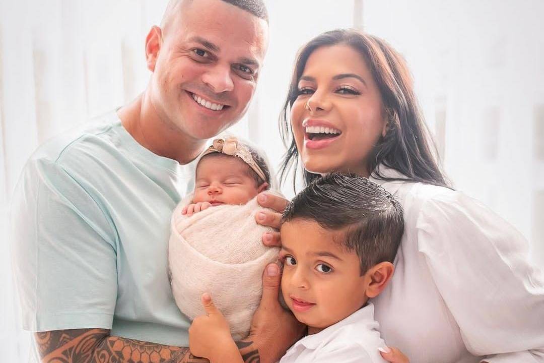 Na imagem, está a família de quatro pessoas: à direita, Evelyn Regly é uma moça morena, de cabelos castanhos e compridosm vestindo camisa branca. À esquerda, seu marido, de cabelo bem curto, camisa azul, segurando uma bebê no colo. A bebê está enrolada em uma manta rosinha, com uma faixinha também rosa na cabeça. À frente da mãe, o filho, menino de quatro anos, camisa branca, cabelo castanho e curto, fazendo sinal de positivo com a mão esqueda.