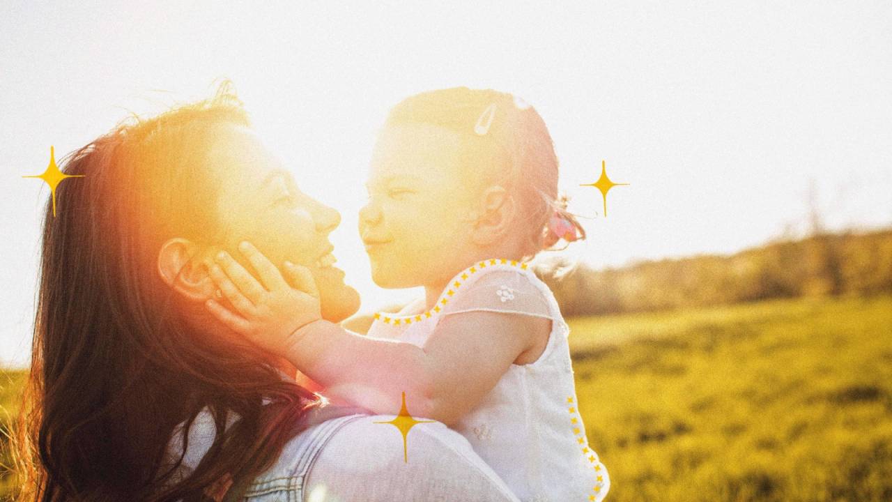 Mulher com menina no colo. Ela tem por volta de 2 ou 3 anos, está com as mãozinhas no rosto da mãe e com o rosto bem próximo do dela. Elas estão no campo, com a luz do sol meio entre elas.