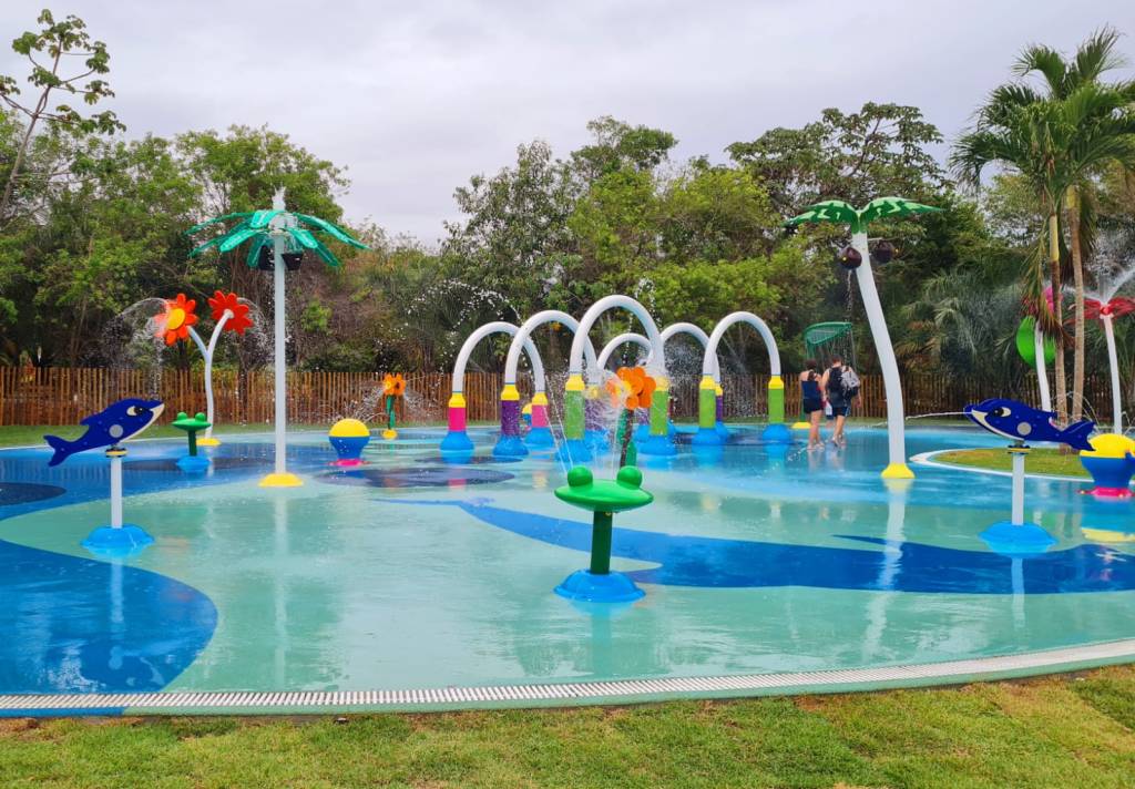 Splash Park Iberostar Na imagem, uma área com sprays de água. O chão sobre o gramado é todo azul e os totens que espirra, água são em formato de flor e de animais marinhos. Ao fundo, árvores e o céu nublado.
