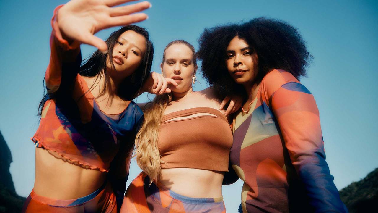 três meninas usando roupas de praia, em um cenário de praia. Elas olham para a câmera