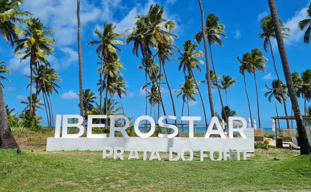 iberostar praia do forte Letreiro branco escrito iberostar praia do forte, sobre hamado. No fundo, céu azul de sol e grandes coqueiros.