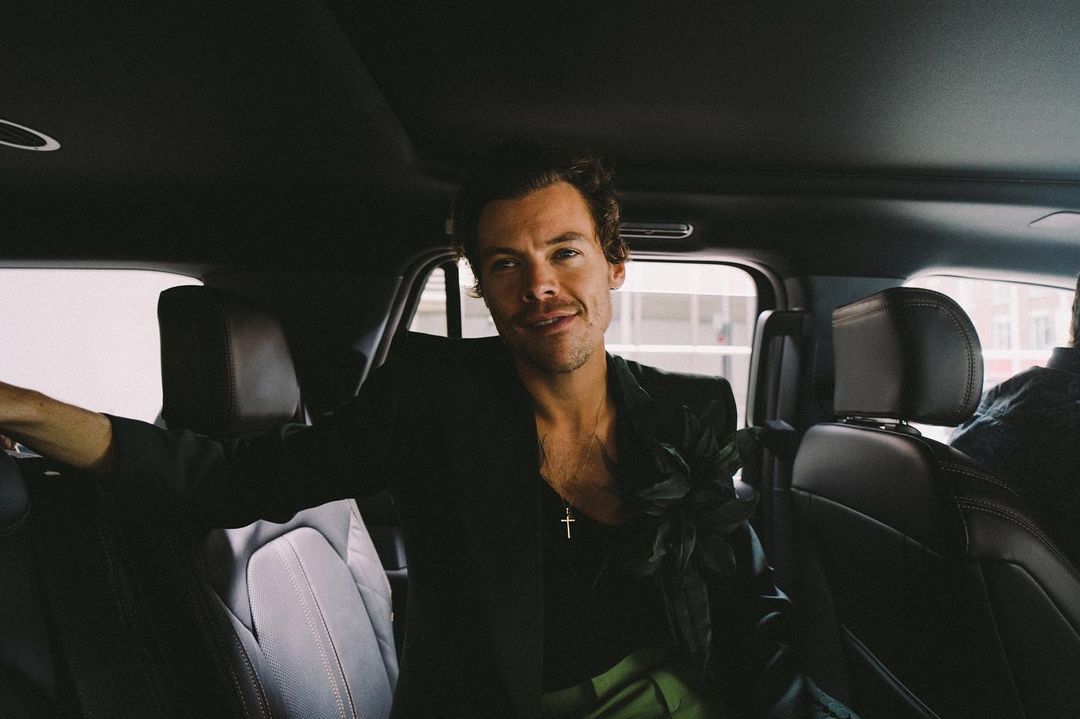 Cantor e atoe Harry Styles dentro de um carro, vestindo uma jaqueta preta e calças verdes